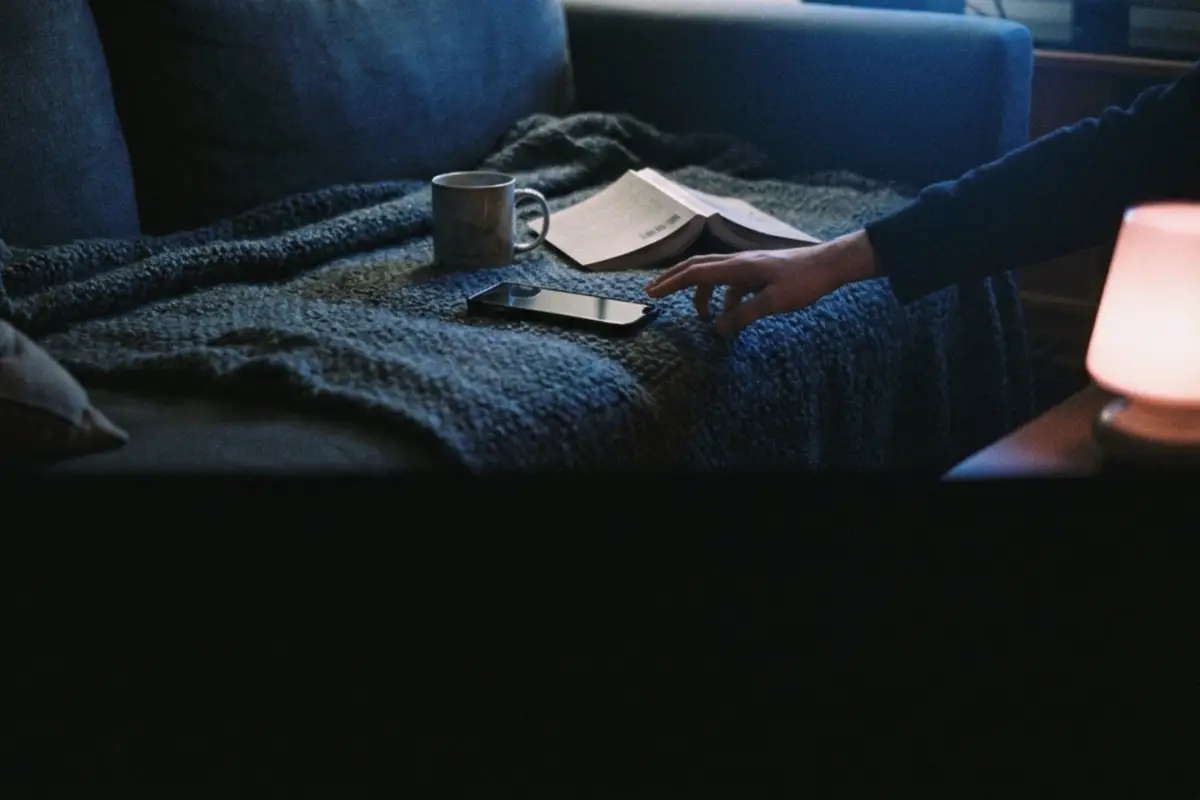 A phone lying dark on a blanket in an unlit living room, a hand reaching for it
