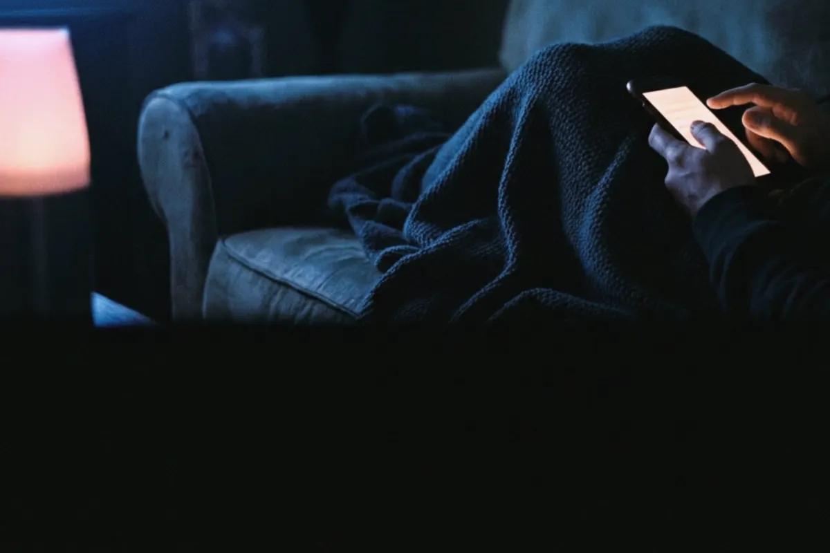Hands cradling a phone in the dark, one thumb mid-tap, a blanket across the lap
